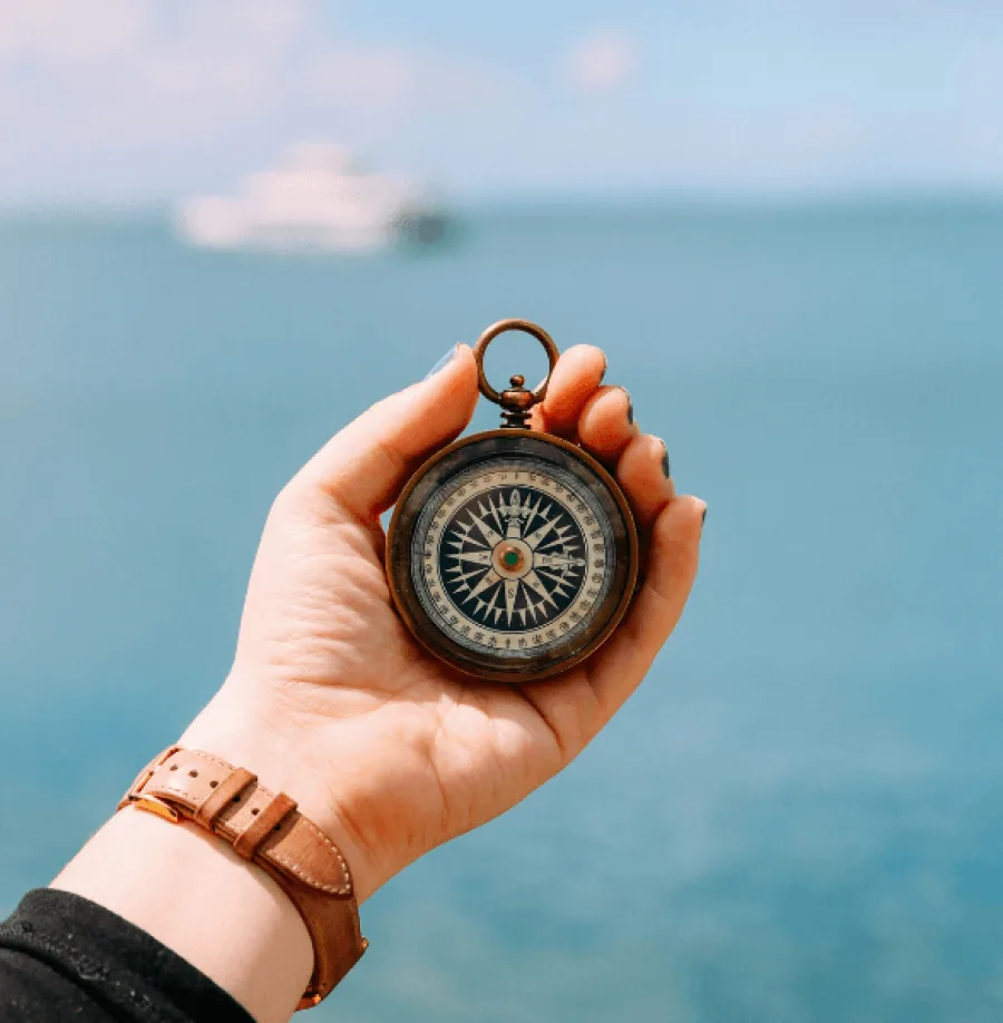Hand hält Kompass vor Meer mit Schiff im Hintergrund