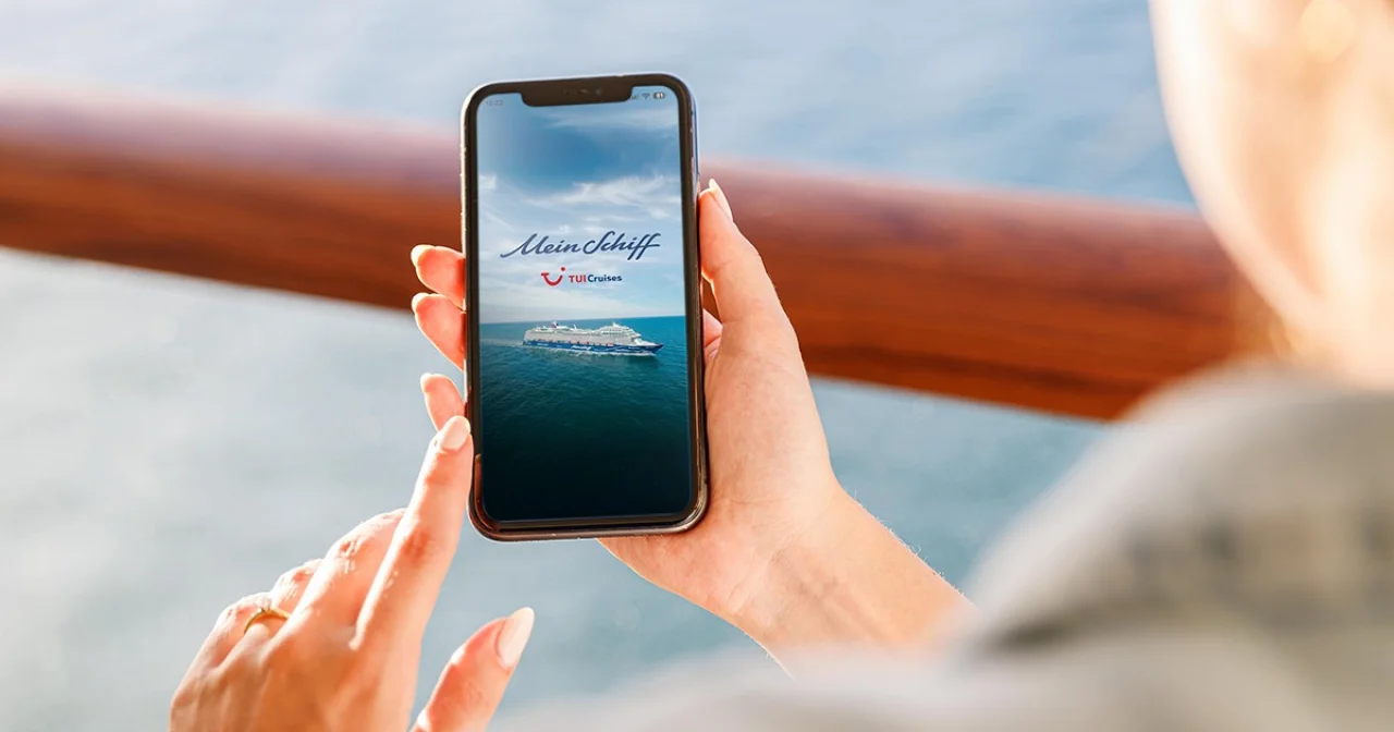 Gast mit Smartphone mit geöffneter Mein Schiff App in der Hand an der Reling eines Kreuzfahrtschiffes