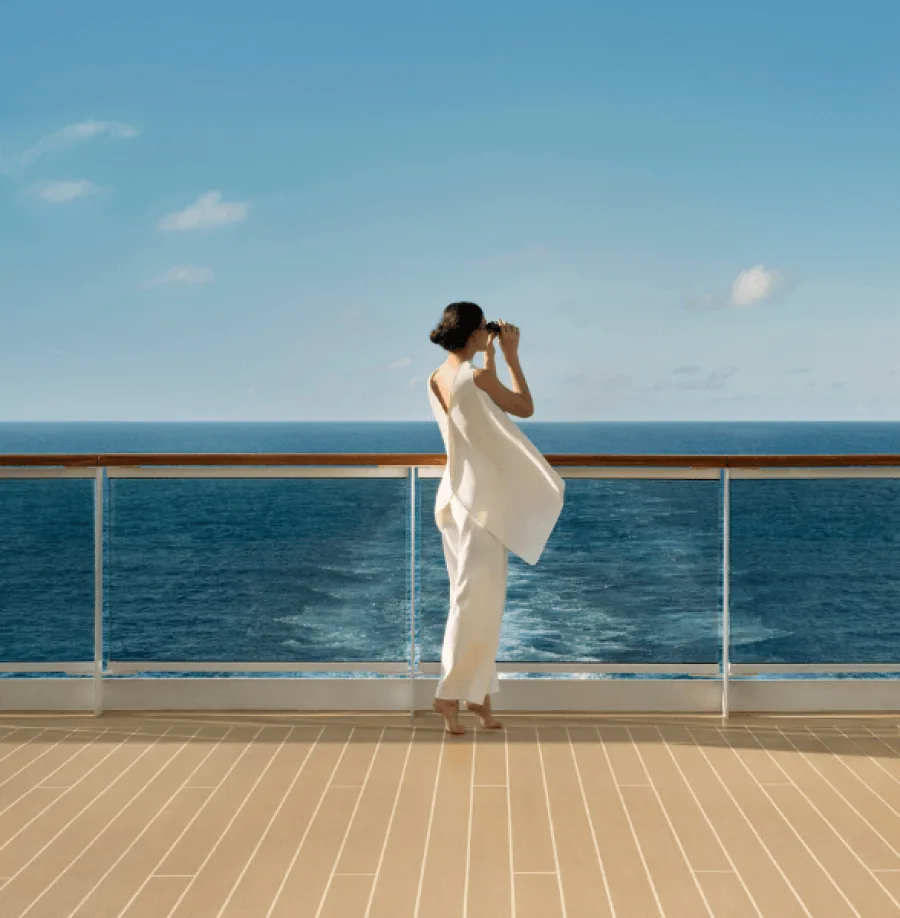 Frau genießt Aussicht mit Fernglas an Reling oberhalb der Heckwelle an Bord eines Kreuzfahrtschiffes von Explora Journeys